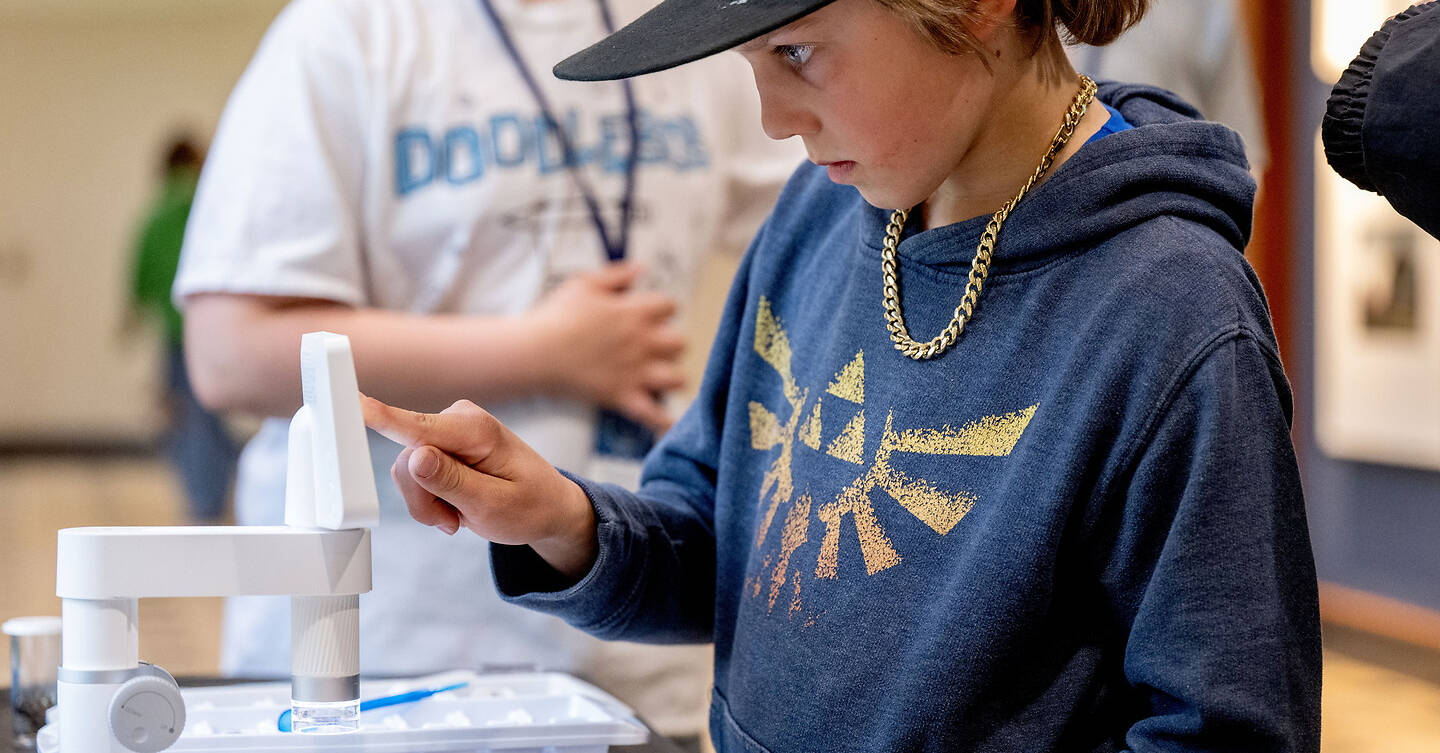 A young k-12 student in a blue hoodie and black cap examines a small white device with a magnifying glass, while others are visible in the background.